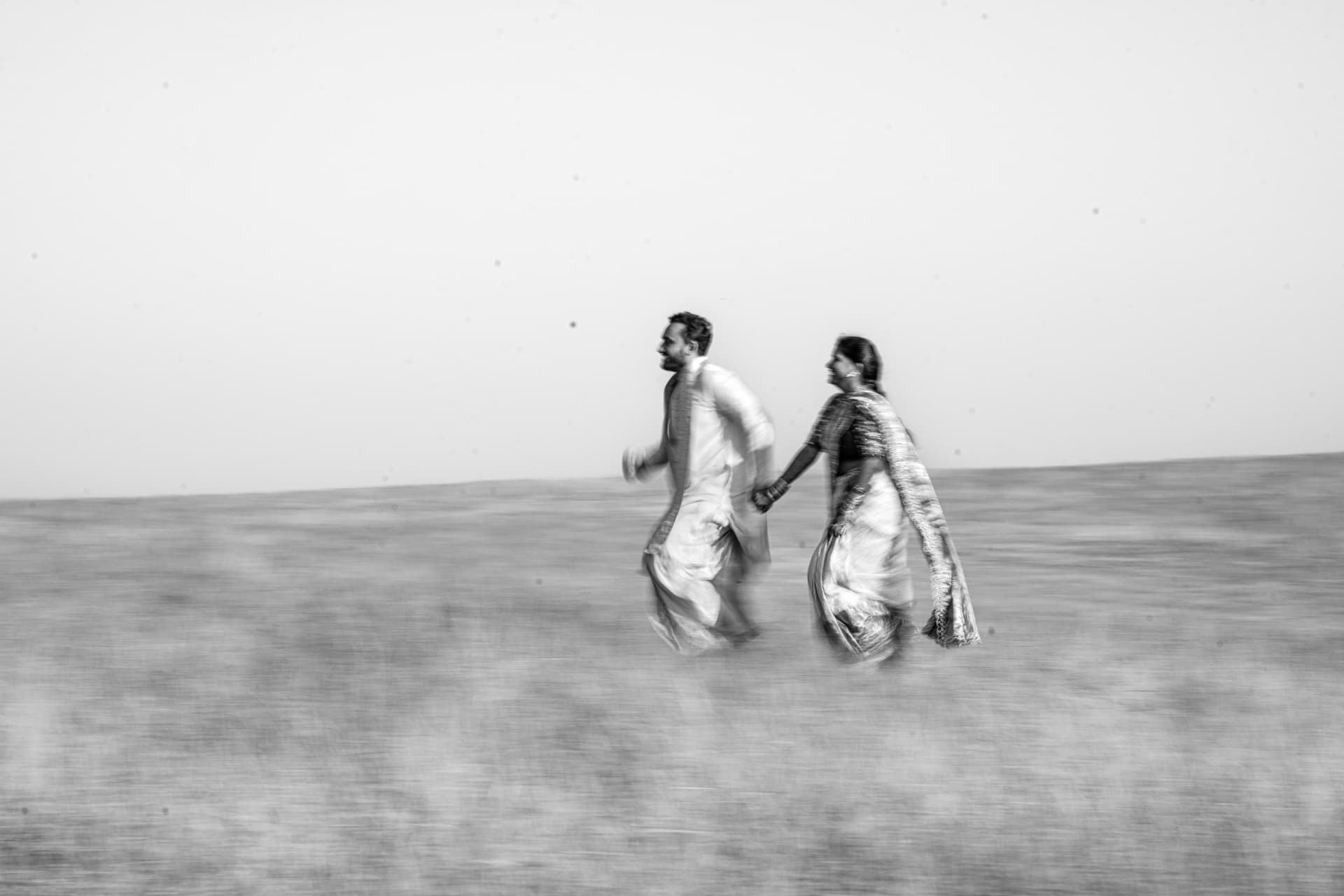 A bride and groom running hand in hand across an open field, photographed in black and white
